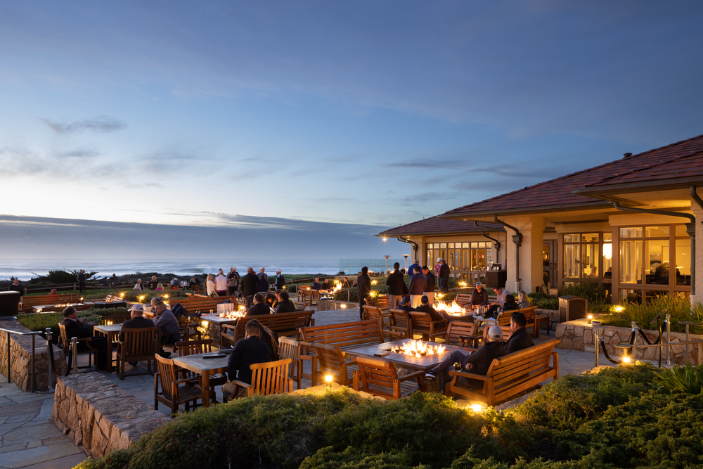 This is an image of the outdoor patio tables at The Inn at Spanish Bay in Pebble Beach, California at dawn. Part of the restaurant can be seen on the right side of the image, with the left side of the image showing several scattered outdoor seating and tables with people at them. The ocean can be seen in the background