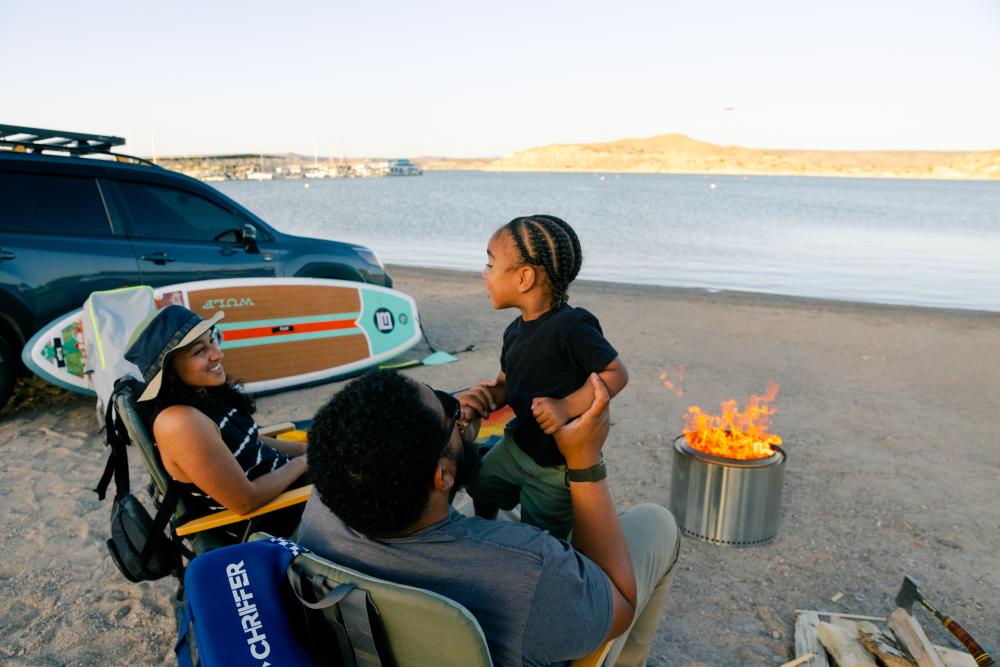 Family gathered around the campfire on the beach of Elephant Butte State Park as the sun sets over the butte and water.