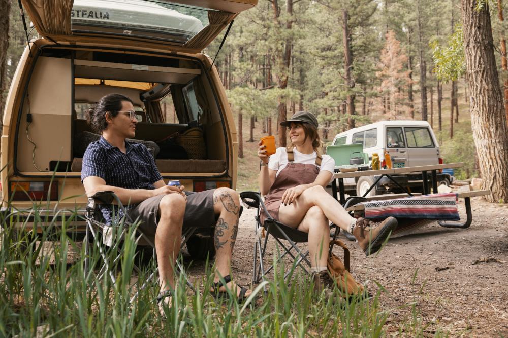 A man and woman relaxing outside of their camper van at a forest campsite in Pecos Canyon State Park, New Mexico.