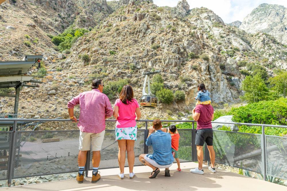 A group of people looking at a tram descending a mountainous terrain, with one adult carrying a child on their shoulders.