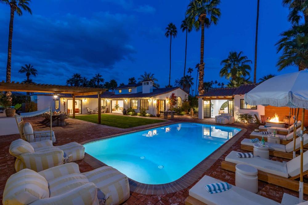 An illuminated luxury backyard with an in-ground swimming pool at twilight, featuring an outdoor lounge area with a fire pit, cushioned chairs, and a covered patio adjacent to a white two-story house surrounded by tall palm trees.