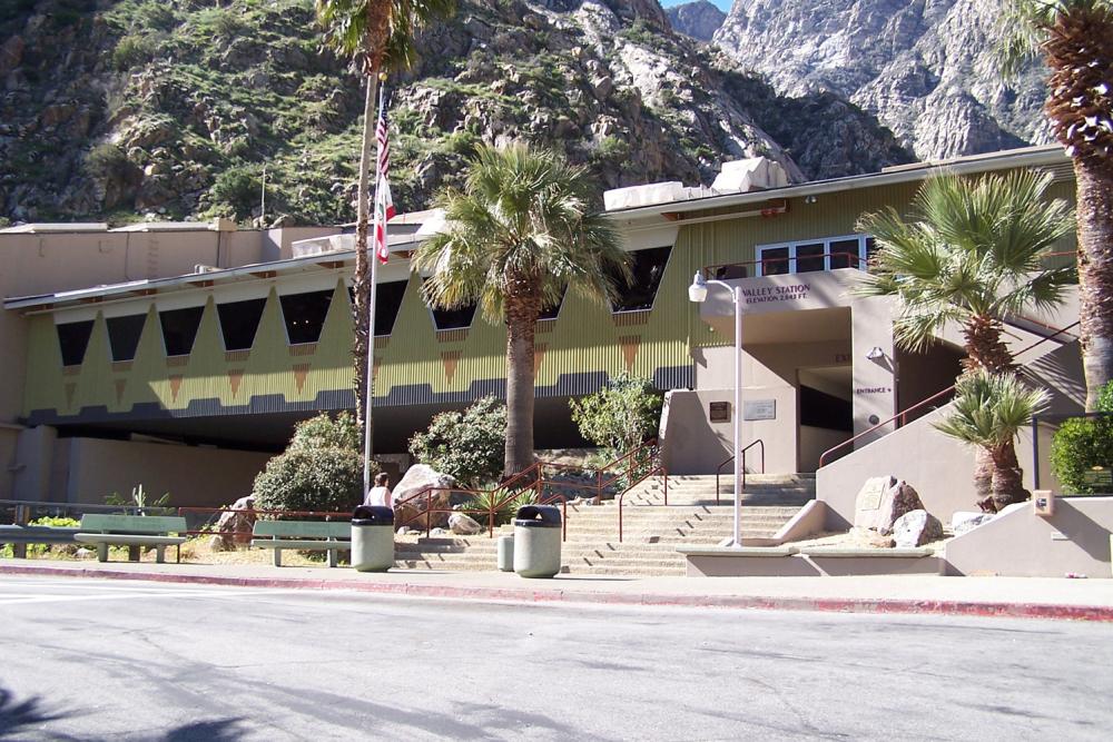 A single-story building with a brown and yellow facade, labeled 'Valley Station' with an American flag and palm trees in front, situated at the base of a rocky mountainside.