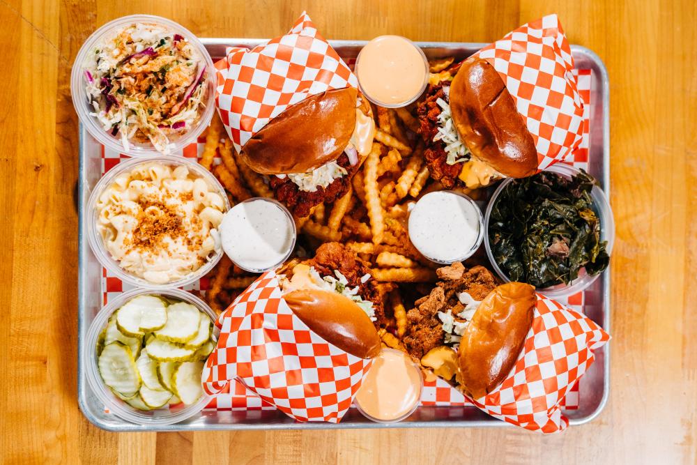 Four Nash and Proper's fried chicken sandwiches on a metal tray on top of fries with sides.