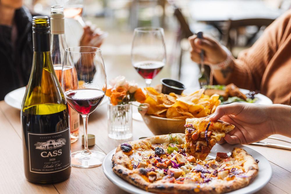 Table with wine bottles, glasses, and food. Person picking up a slice of pizza.