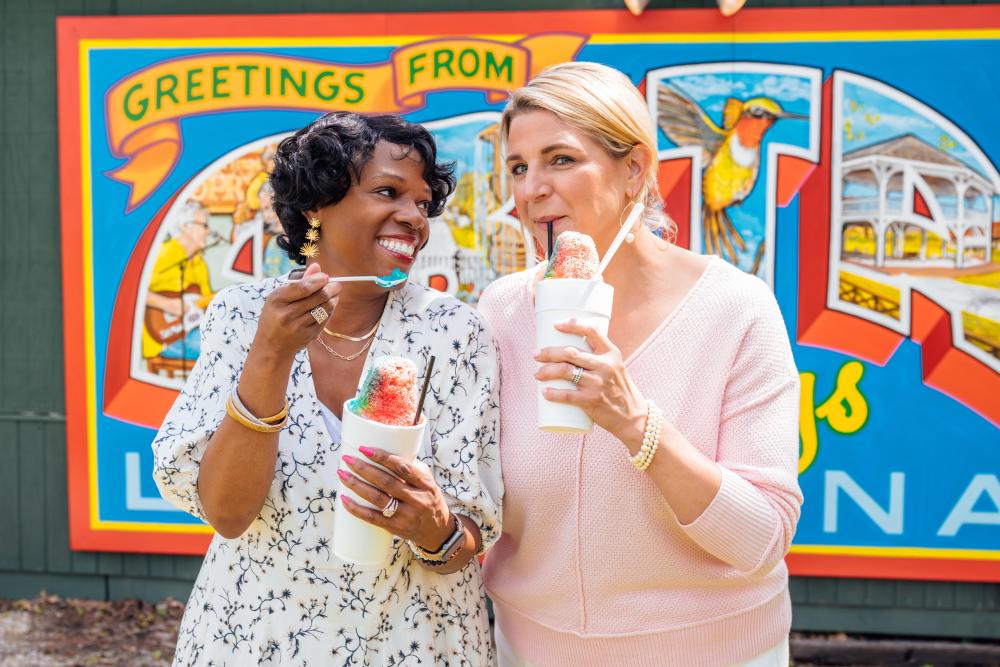 Zondra White Jones and Katie Guasco enjoy colorful snowballs from The Stand in Abita Springs while standing in front of the shop’s vibrant mural. The mural, painted in a vintage postcard style, features local landmarks, creating a colorful backdrop as the two podcast hosts smile and share a moment together outdoors.