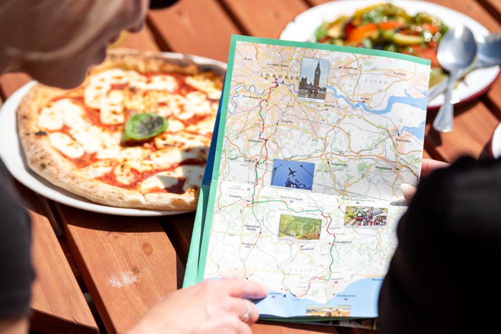 Looking at the Avenue Verte Map, a paper map in focus, outside with a pizza on the table