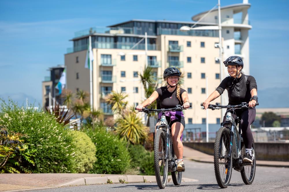 two ladies cycling on a road in town with a nautical building behind them.