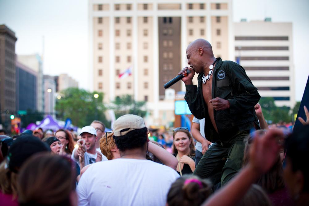 Lead singer for Fishbone screams into a mic in front of a large crowd on an outdoor stage at Wichita Riverfest