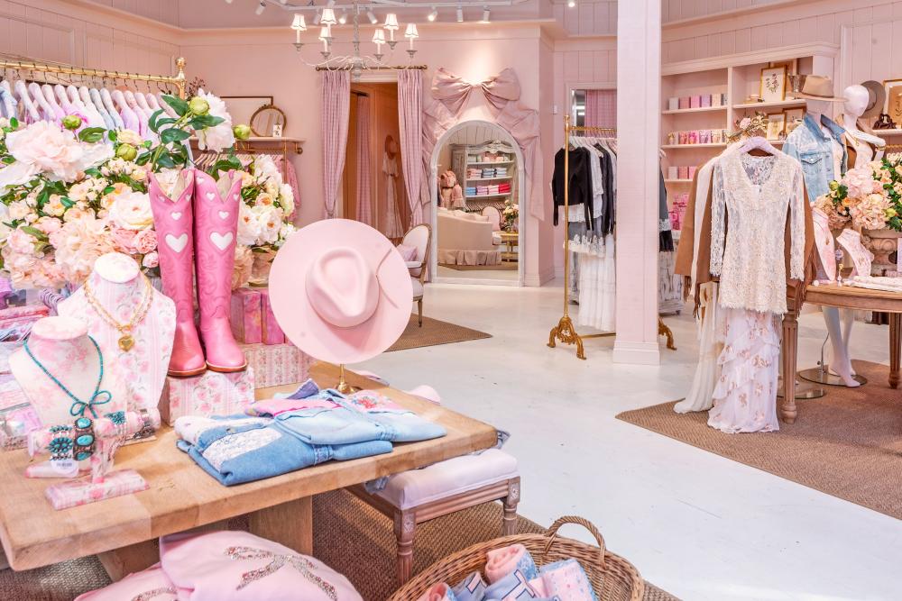 The interior of LoveShackFancy is all things pink. On the table nearest the viewer are bright pink cowgirl boots with white heart cutouts, a pale pink cowgirl hat, blue jeans with pink floral patches over the pockets, etc. All walls and decor, and most products, are some shade of light pink.