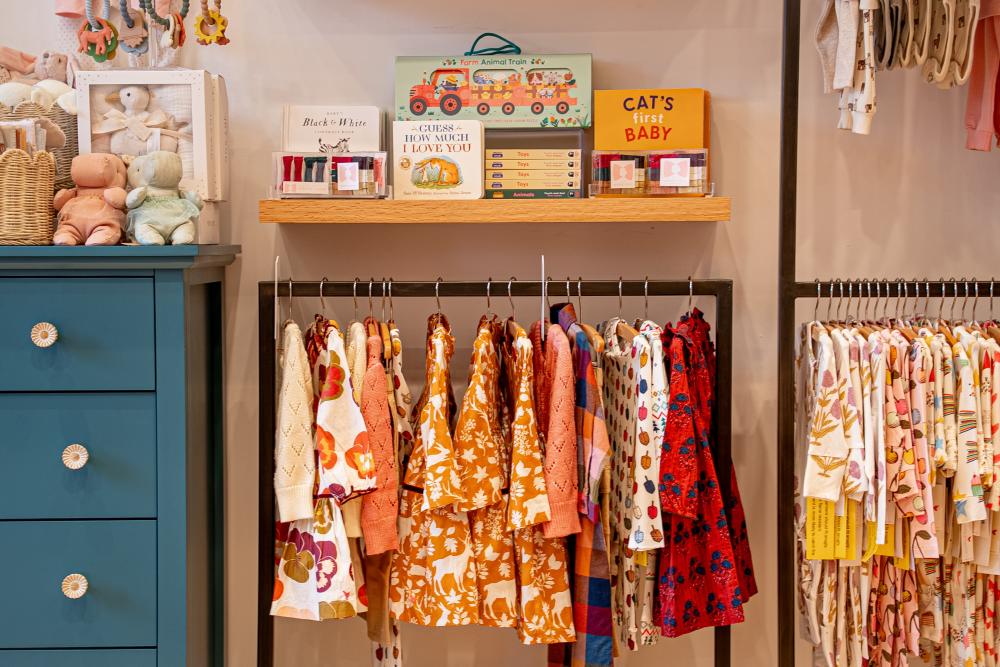 A display at Pink Chicken. On the left, a blue dresser topped with plush hippos and soft teething rings. In the center, a clothes rack with floral fall dresses and a shelf lined with baby books. On the right, a rack with colorful kids' pajamas.