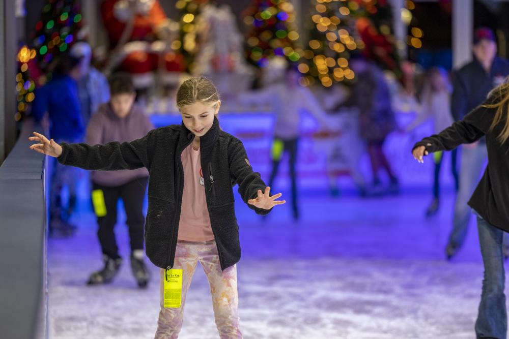 A tween girl skates solo around The Woodlands Ice Rink. She's dressed in pink casual clothes and a fuzzy black jacket.