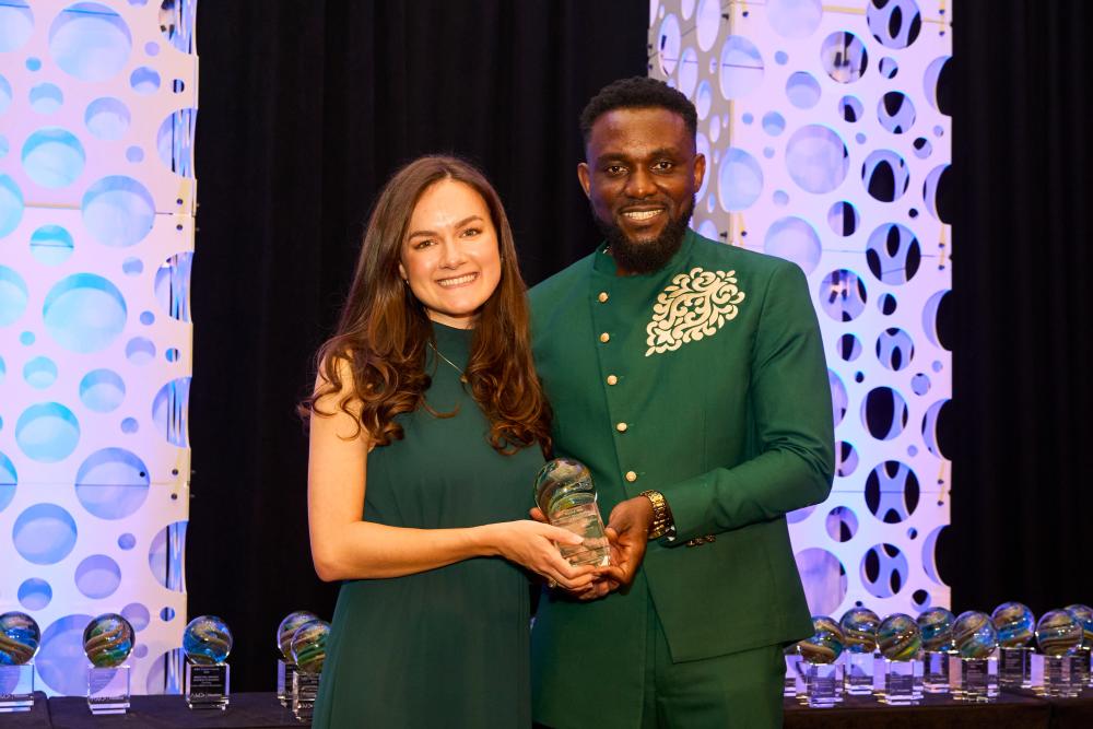 The MC of the 2026 AMA Crystal Awards poses for a photo with Andrea Day, winner of the Advertising-Photography category. Andrea is wearing an elegant dark green dress. The MC is wearing a forest green suit with silver decals. They're holding a decorative glass globe. Decorative hole-filled pillars lit in blue and purple stand behind them.