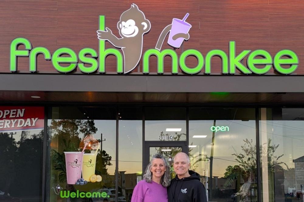 An older couple, Crispin and Mitch Provost, stands in front of their new Fresh Monkee franchise. Crispin has long salt-and-pepper hair and a bright lilac hoodie. Mitch has little hair, all gray, and is wearing a black hoodie.