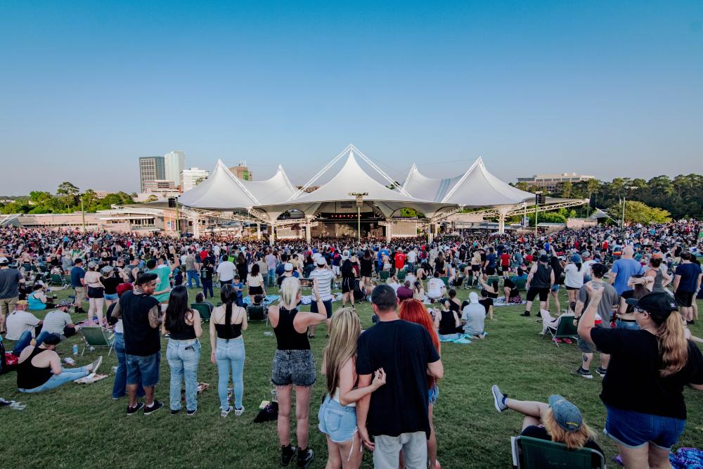 An afternoon photo of The Pavilion, viewed from the back of the lawn. The lawn and seats are packed with a crowd of young adults, all dressed casually for summer. The Pavilion tents rise in 5 points, the stage lit by the afternoon sun. The Woodlands Towers are partially visible behind it.a crowd of casually dressed 20- and 30-year-olds