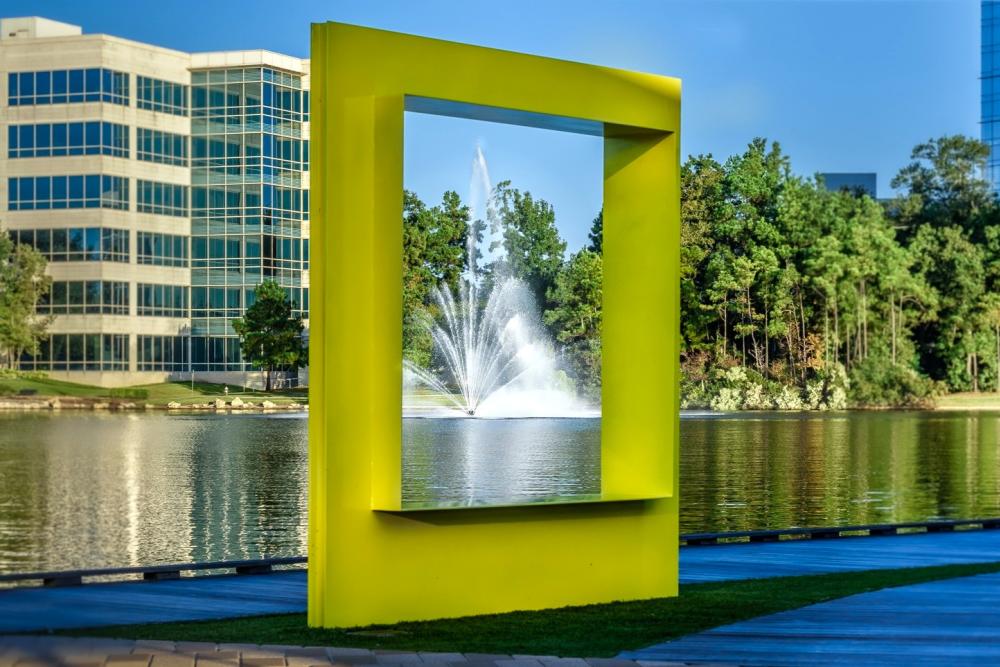 The Woodlands Wind-O is a large yellow sculpture shaped like a picture frame or a window overlooking Lake Woodlands at Hughes Landing.