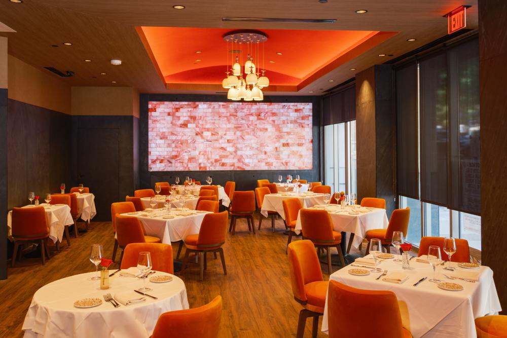 The main dining room at Bari Ristorante at Waterway Square has light wood floors and 10+ square tables with plush orange seats. Each table is set for fine dining.