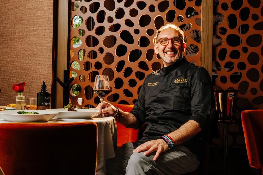 Bari Ristorante's Master Chef Renato de Pirro sits at a table in the main dining room. The table set with three gourmet dishes and a glass of wine.
