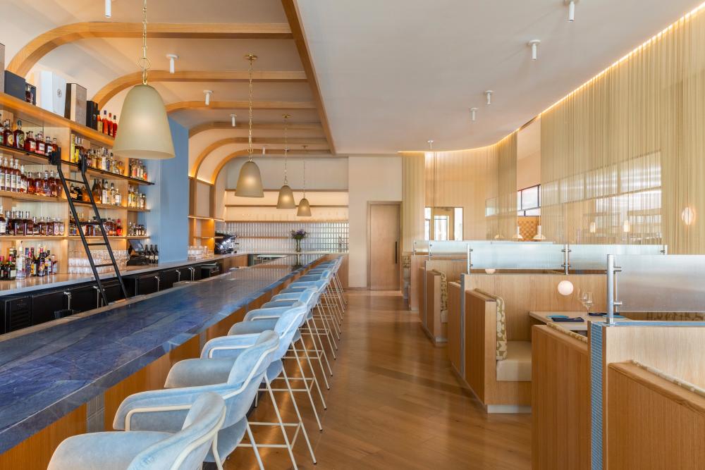 The bar at Charolais by Chef Austin Simmons features a denim-blue countertop and plush light blue chairs. The walls, floors, and booths along the right wall are all made of light wood and tan fabrics.
