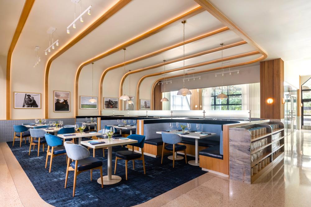The dining of Charolais by Chef Austin Simmons features blue carpet, thin white tables for two, plush blue chairs, and fine cutlery. Wall decor consists mostly of light wood frames on Western photography.