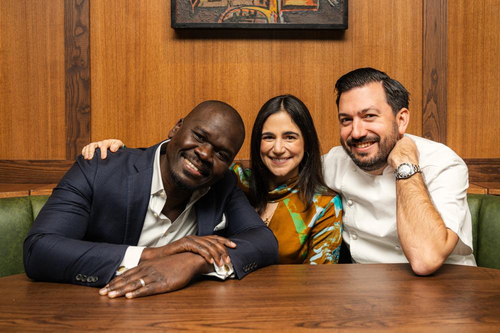 Three people sit at a Bar Bludorn table, smiling at the camera. From left: Cherif Mbodjij, Victoria Bludorn, and Aaron Bludorn. They are the co-founders of Bludorn Hospitality Group.