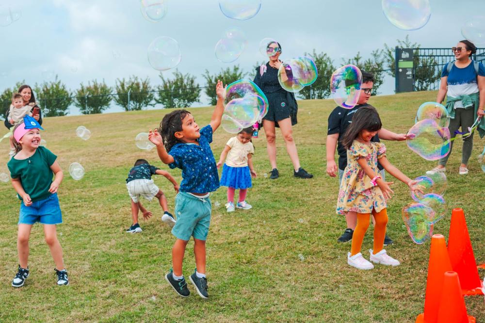 A group of young children reach up to pop large bubbles floating across The Lawn at The Pavilion during the 2025 children's festival.