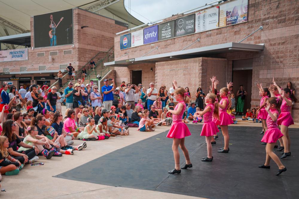 A group of young girls in pink leotards dances in the plaza at The Pavilion during the 2025 Children's Festival. Other kids and their families sit on the ground around them to watch.