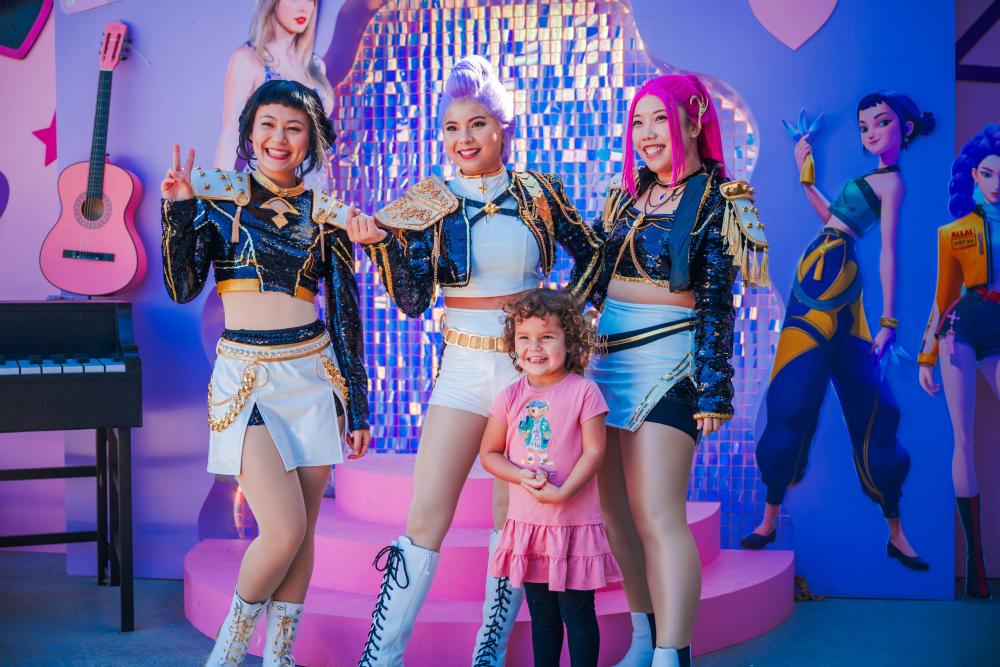A young girl in a pink dress smiles for the camera during a character meet at The Pavilion's Children's Festival 2025. The characters are Zoey, Rumi, and Mira from the Netflix film, "Kpop Demon Hunters."