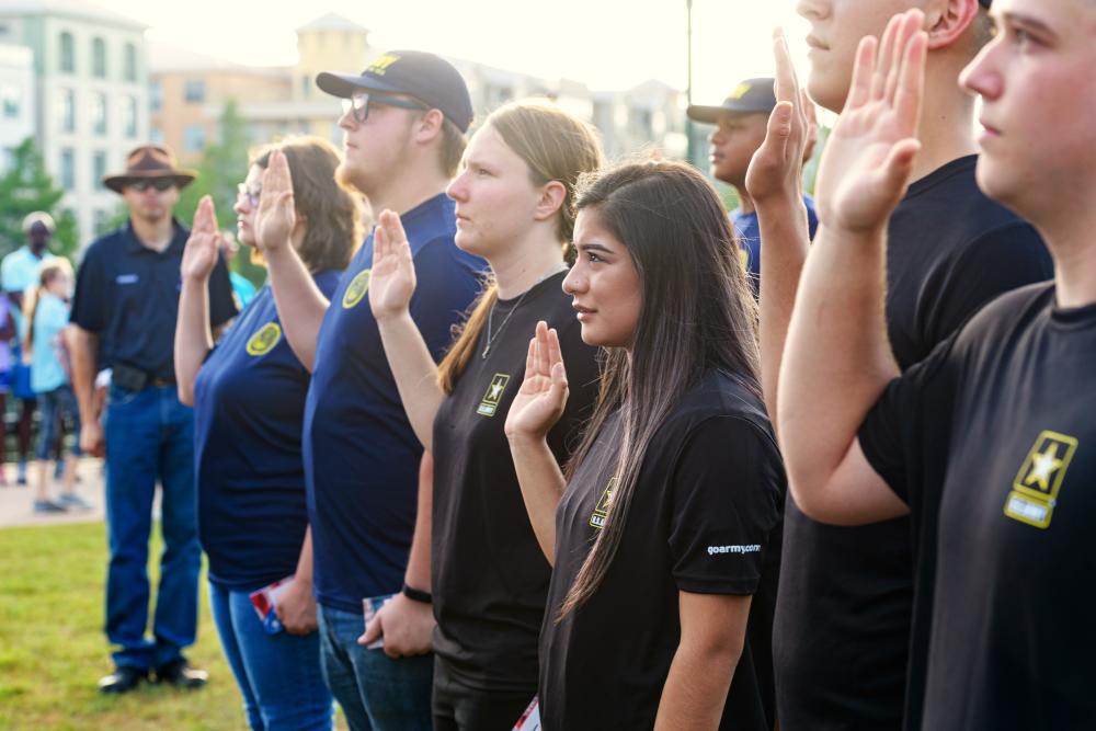 A group of young men and women are sworn in to the U.S. military. The camera focuses on a row of four initiates (three women, one man) raising their right hands to be sworn in.