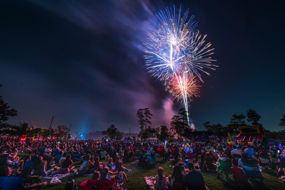 A bouquet of red, white, and blue fireworks burst in the night sky over Town Green Park. Hundreds of spectators stare up at the spectacle. Along the horizon are dark silhouettes of trees and a large, inflatable water slide.