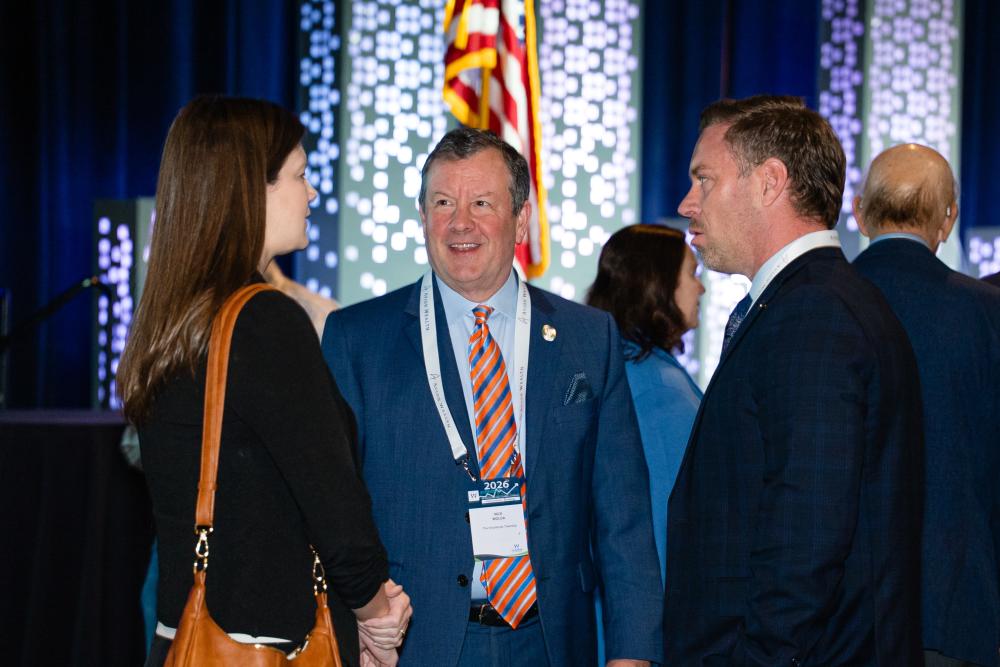 Nick Wolda (blue suit with an orange-and-blue striped tie) converses with a woman in a dark suit with a brown purse and a man in a dark suit.