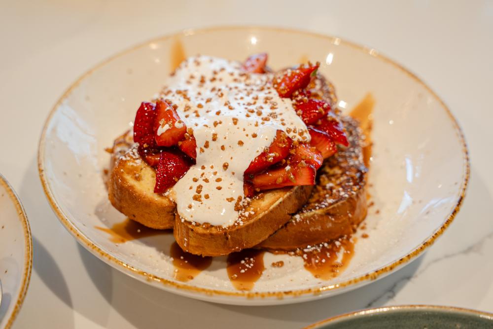 A shallow bowl is filled with French toast topped with syrup, cream, and fresh chopped strawberries.
