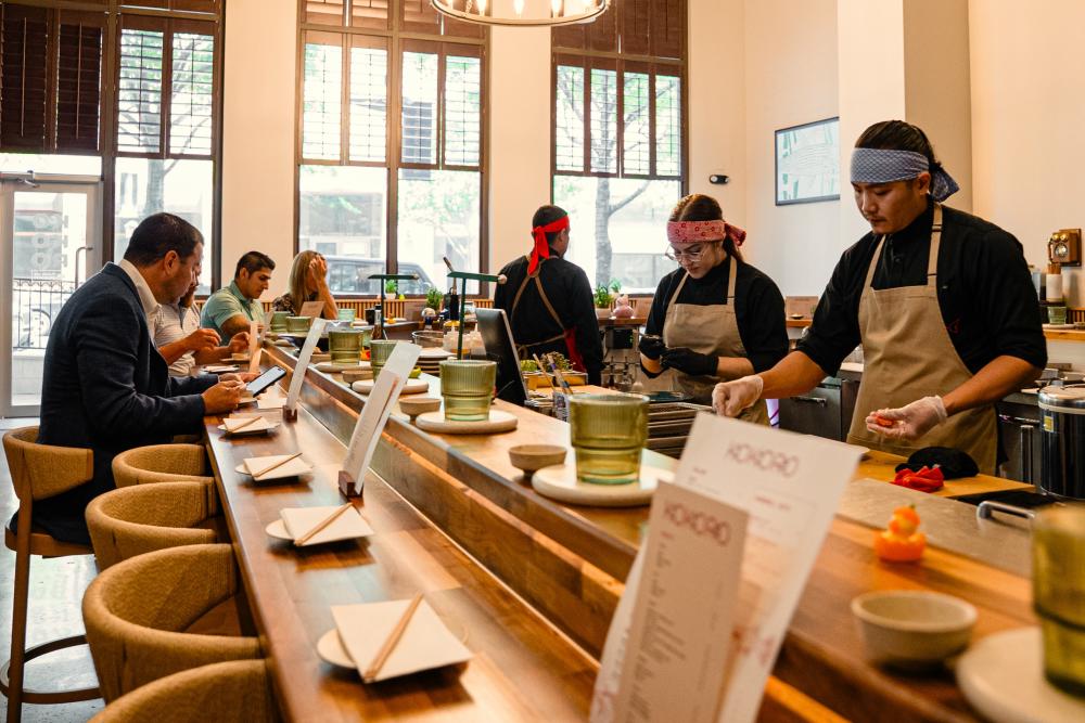 A view of the Kokoro handroll bar from the back of the dining room. Three chefs prepare dishes along the inside of the bar; four guests are seated on the opposite side. Light pours in from tall windows in the front.
