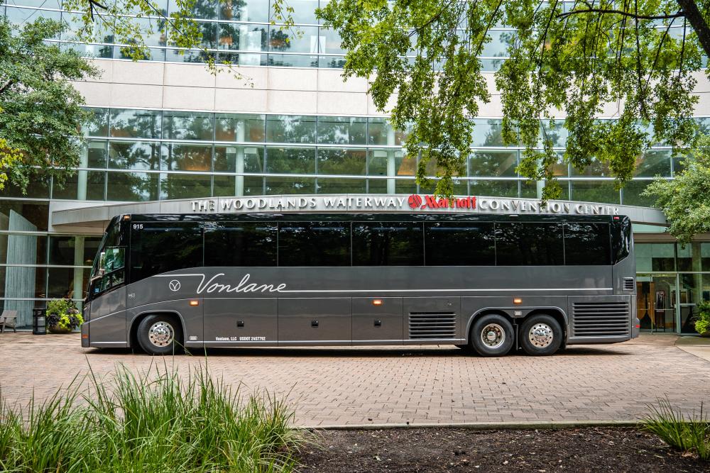 A long silver bus bearing the "Vonlane" name and logo is parked in front of The Woodlands Waterway Marriott Hotel & Convention Center. The front of the hotel is all glass windows, and greenery frames the image.