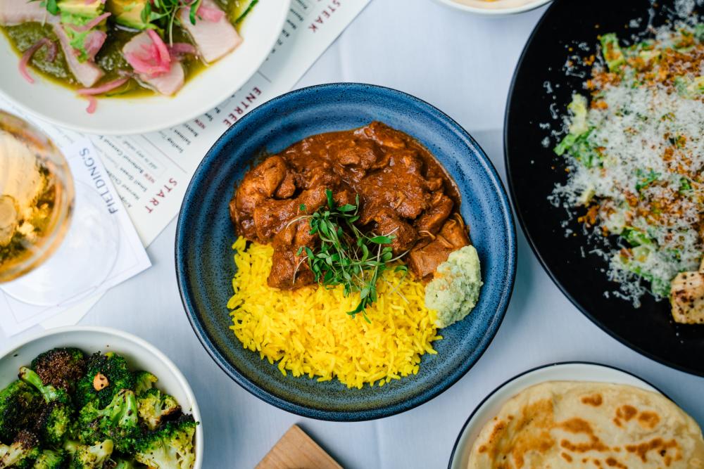 An overhead view of a lunch spread at Fielding's Local Kitchen + Bar. The central dish is Vegetable Coconut Curry Masala, featuring chicken in a thick reddish sauce, yellow rice, and herbs in a bright blue bowl. Around it are a tuna poke bowl, a chicken salad, naan bread, and hazelnut broccoli.