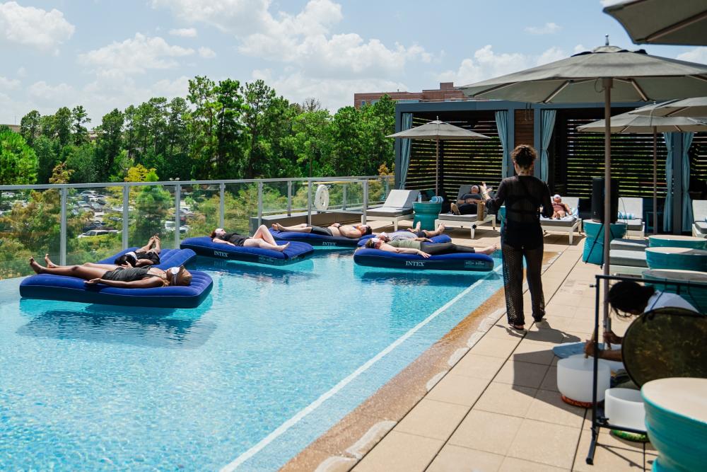 Six people relax on inflatable mattresses in the rooftop infinity pool at The Westin at The Woodlands. Beside the pool, a woman in black offers therapeutic words while a man plays singing quartz bowls, wind chimes, and a gong.