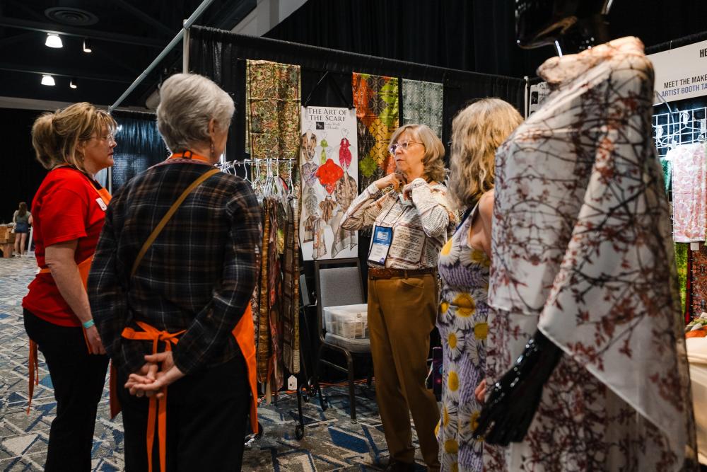 Artist Heather Wasaff demonstrates methods for wearing scarves she created. Three women of various ages listen intently at her booth during the Texas Fine Craft Show.