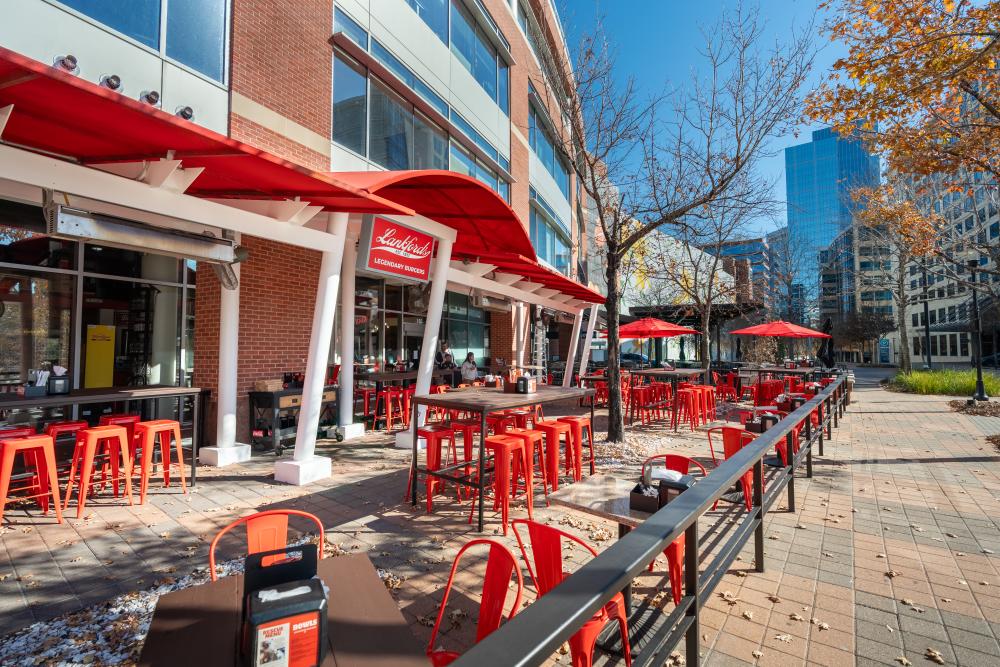 The exterior of Lankford's is a brick building with a bold red and white color scheme. A long patio features at least a dozen thick wooden tables with bright red chairs or bar stools. The Waterway Square Parking Garage and The Westin are visible in the background.