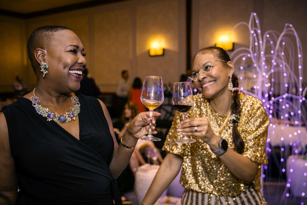 Two black women wearing gold sequins and lavish jewelry toast with red and white wine at the W&FW Grand Tasting.