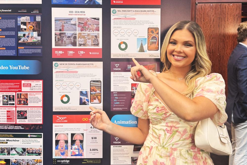 Brooklyn Newell poses with two of her AMA Crystal Award entries that became finalists. She has dark hair with blonde highlights and is wearing a light pink-and-white floral dress. She's pointing to her poster submissions: "No, This Isn't a Hallmark Movie..." and "New In Town: Paris Baguette."