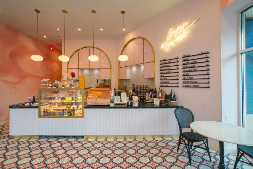 The interior of Kendra Scott Sips & Sweets is very pink. One wall is pale pink with gold accents, another is a marbled swirl of various pinks. The flooring is a mosaic of repeating tile patterns in pink, white, black, and blue. Drink options are listed in big black text on the wall.