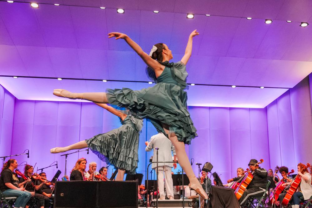 Two ballerinas in teal dresses perform elaborate jumps on the main stage of The Cynthia Woods Mitchell Pavilion. A full orchestra plays behind them. The ceiling and walls are lit with blue and purple lights.
