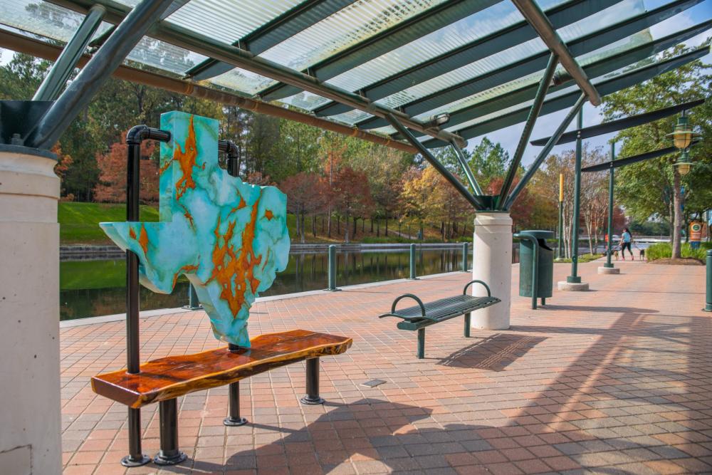 The State of Texas art bench features a large slab of turquoise (shaped like the state of Texas) marbled with copper inlays. It stands upright on a smooth wooden bench, propped up by black metal pipes. It is located along The Woodlands Waterway behind The Pavilion.