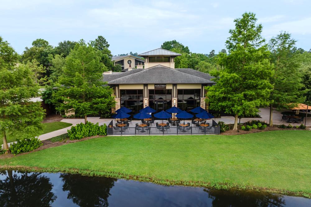 An exterior shot of Back Table Kitchen & Bar at The Woodlands Resort. It's a cream-colored building supported by four columns. Nine wooden tables with deep blue umbrellas line the patio beside the golf course.