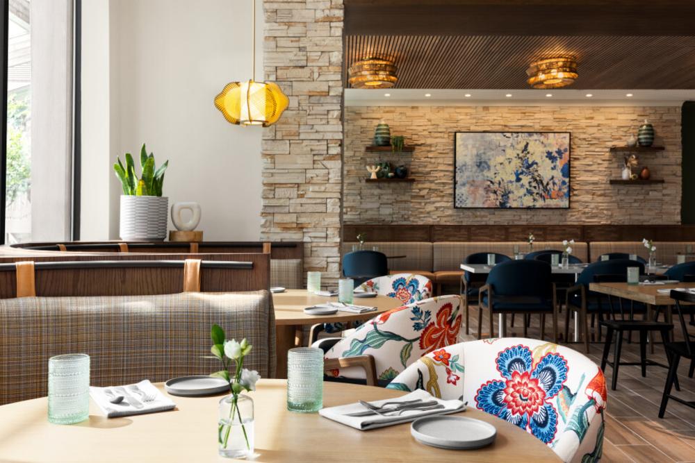 A close-up view of a dining room at Harrison's, a lakeside restaurant at The Woodlands Resort. The room features a number of round tables with bright, floral-patterned chairs and square tables with dark blue chairs. There's a window on the left revealing golden hour sunlight.