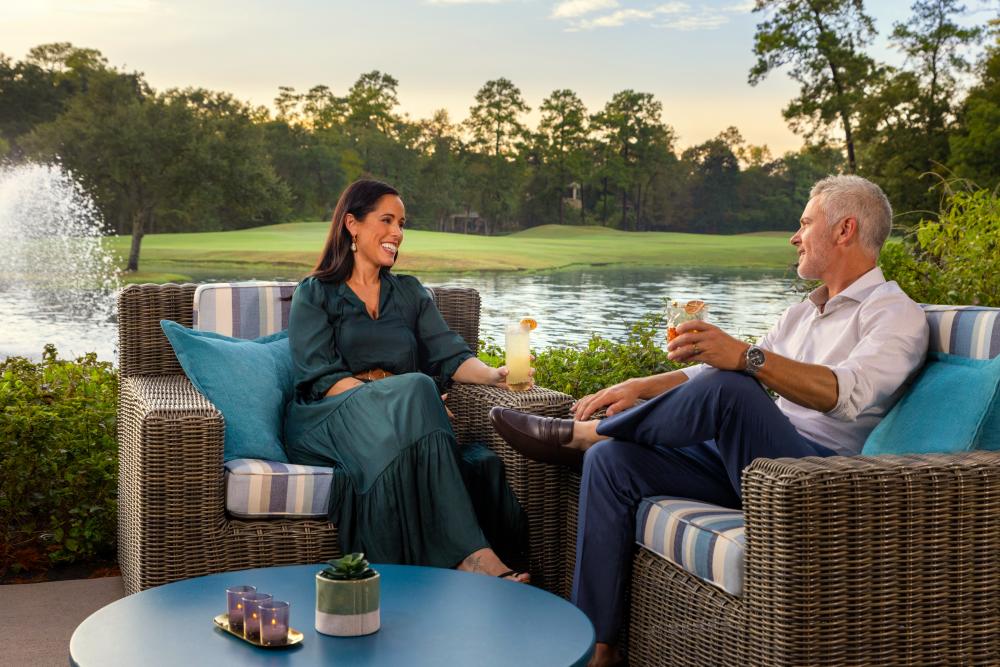 A couple enjoys cocktails at sunset on a patio of The Woodlands Resort. They're sitting in large whicker chairs with blue-striped cushions. The patio overlooks the golf course.