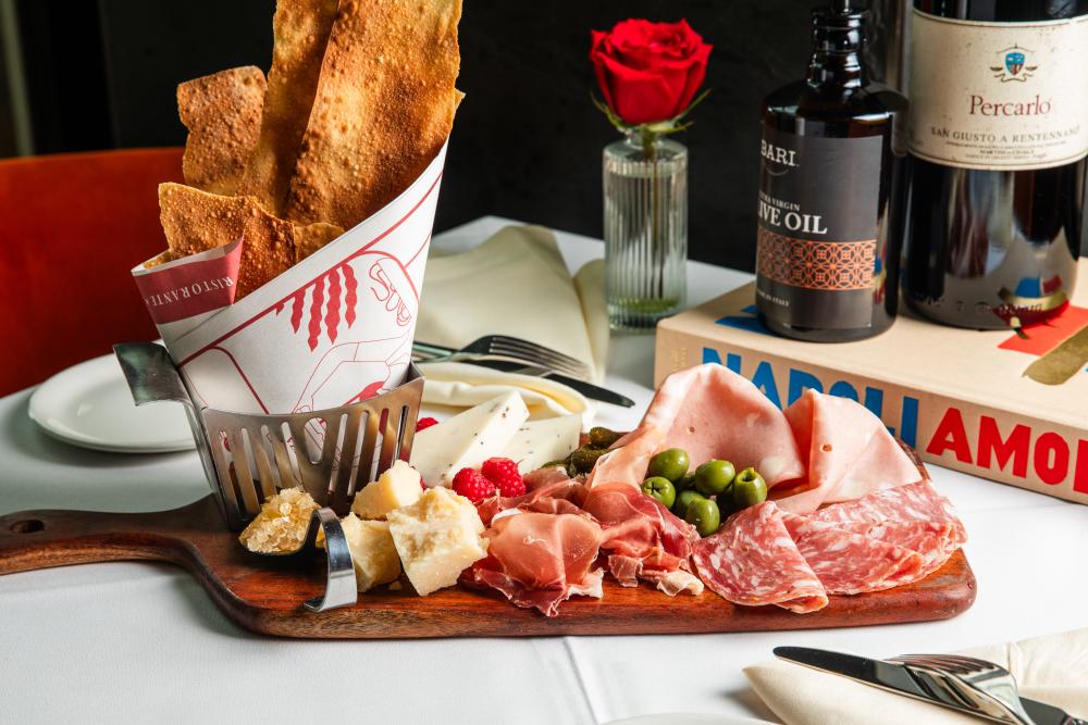 A charcuterie board is set with various gourmet meats, cheese chunks, green olives, pickles, and raspberries, served with a basked of crisps.