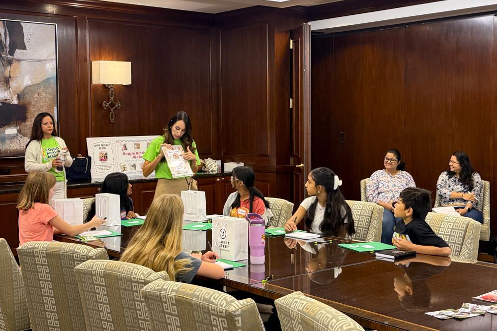 Noemi Gonzalez teaches a workshop during the 2026 Young Makers Market. A diverse group of 6-8 students (approx. 8-10 years old) listens intently. The workshop is set in a fancy conference room with dark wood walls and plush chairs. Some of the children's parents observe from the doorway.