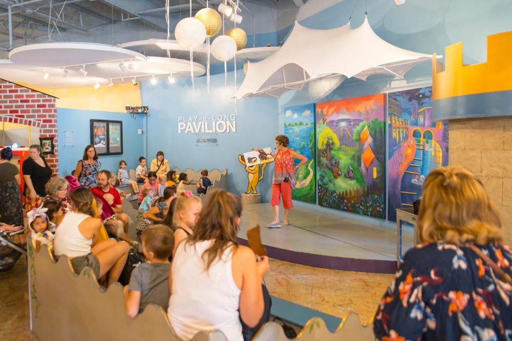 Inside TWCM is a stage set up to look like The Cynthia Woods Mitchell Pavilion, complete with tent awning and a tri-paneled mural with iconic landmarks around The Woodlands. The audience is full of families with young kids, all listening to a woman reading a children's book on the stage.