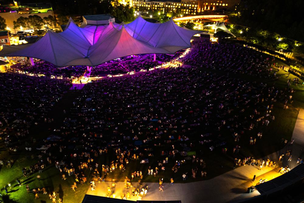 An aerial drone view of The Pavilion at night. The tents are lit pink and purple, the crowd partially lit with soft yellow lights.