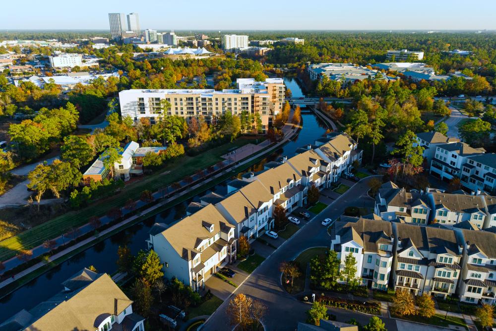 An aerial drone view of The Woodlands during late afternoon. The camera hovers over residences along the south/west arm of The Waterway, facing The Pavilion and The Woodlands Towers.